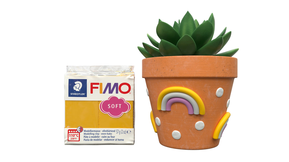 A package of FIMO Soft clay next to a terracotta pot decorated with a rainbow and white dots, topped with a green succulent, on a white background.