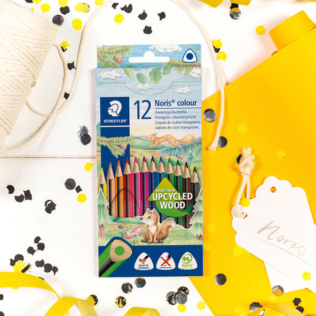 A package of 12 Noris Colour drawing pencils by Staedtler, displayed on a white surface with decorative yellow ribbons, black and yellow confetti, a spool of thread, and a yellow gift bag.