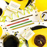 A styled flat-lay birthday party scene with yellow balloons, black plates, yellow streamers, cupcakes, coffee cups, confetti, and several colored pencils on a white surface.