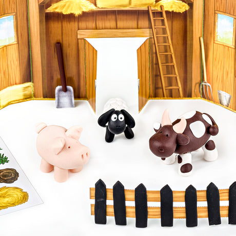 Fimo small farm-themed toy figurines—a pink pig, a black sheep, and a brown-and-white cow—placed on a white surface in front of a miniature wooden barn scene with a fence in the foreground.