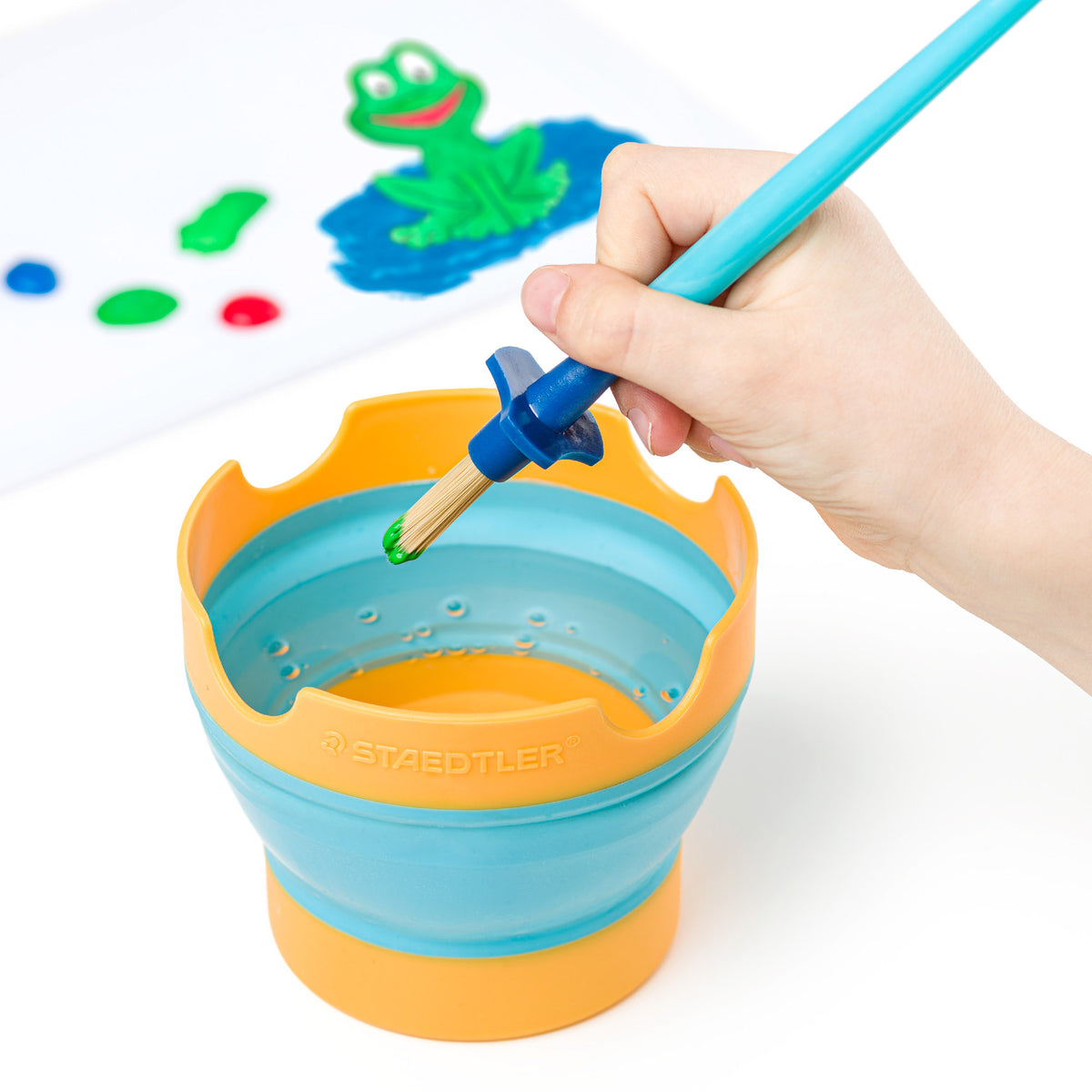 A hand holds a paintbrush over a Stäedtler collapsible paint bucket, which is orange and light blue. In the background, a white sheet shows a green frog painting and paint splatters.