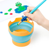 A hand holds a paintbrush over a Stäedtler collapsible paint bucket, which is orange and light blue. In the background, a white sheet shows a green frog painting and paint splatters.