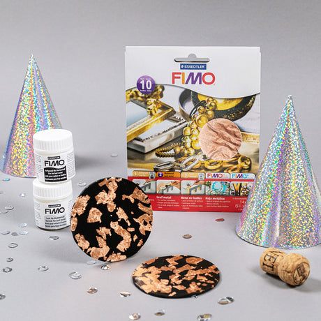 Fimo clay craft kit with a white retail box, two white bottles of Fimo clay, two iridescent silver party hats, two round black-and-rose-gold patterned discs, a cork, and scattered sequins on a light gray background.