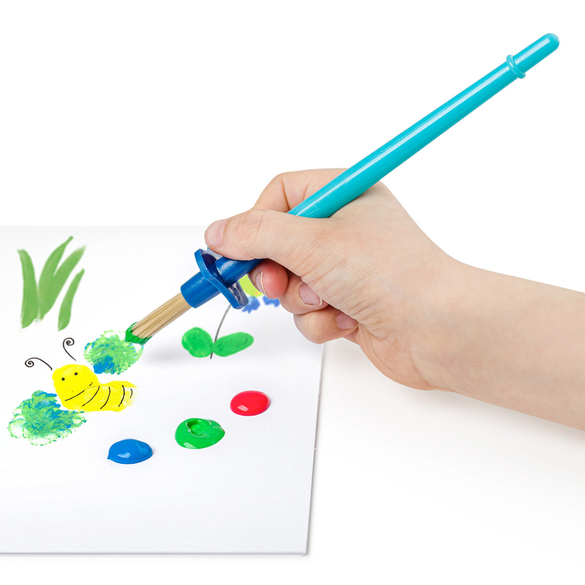 A hand holding a turquoise and blue paintbrush with natural bristles, painting on a white sheet of paper. Drops of green, blue, and red paint, along with a yellow painted caterpillar, are visible on the paper.