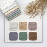 Six square fimo clays in neutral and earthy tones are arranged on a white board, with a matching stack of folded swatches in the top left and dried floral stems in the top right. The swatches are labeled with names including Seabird, Sunflower, Mist, Mallow, Spinach, and Rose Hip.