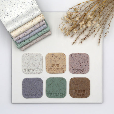 Six square fimo clays in neutral and earthy tones are arranged on a white board, with a matching stack of folded swatches in the top left and dried floral stems in the top right. The swatches are labeled with names including Seabird, Sunflower, Mist, Mallow, Spinach, and Rose Hip.