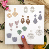 Assorted earrings on a white card held by a hand with a purple nail, surrounded by flowers and leaves.