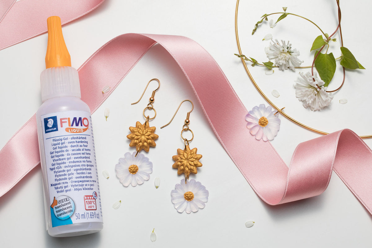 A bottle of FIMO Clay Master polymer clay with an orange nozzle, styled on a white background with a pink ribbon, gold daisy earrings, and white flowers.