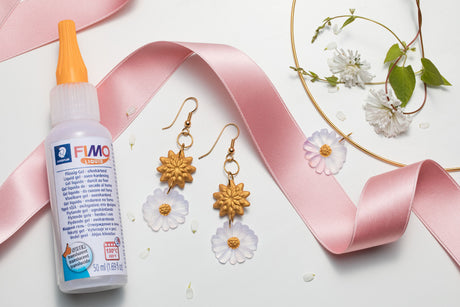 A bottle of FIMO Clay Master polymer clay with an orange nozzle, styled on a white background with a pink ribbon, gold daisy earrings, and white flowers.