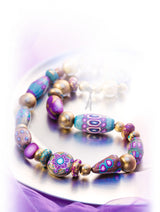 A Fimo Professional Polymer Clay beaded bracelet with colourful patterned beads in purple, blue, teal, and metallic gold, displayed on a reflective silver plate with a soft purple glow.