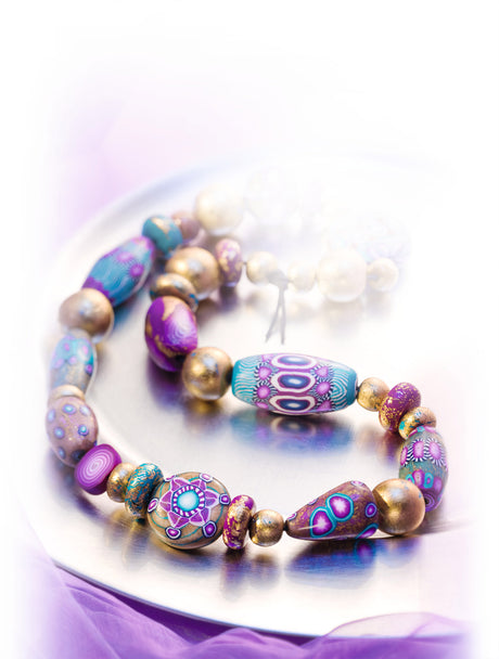 A Fimo Professional Polymer Clay beaded bracelet with colourful patterned beads in purple, blue, teal, and metallic gold, displayed on a reflective silver plate with a soft purple glow.