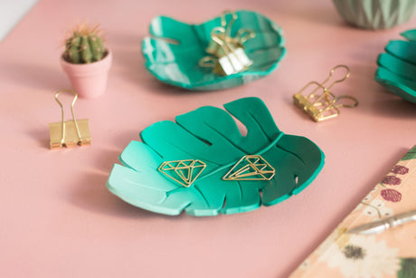 A styled flat-lay featuring two teal and green gradient monstera leaf-shaped fimo polymer clay trays holding gold wire diamond clipboards, alongside other gold paperclips on a pink background.