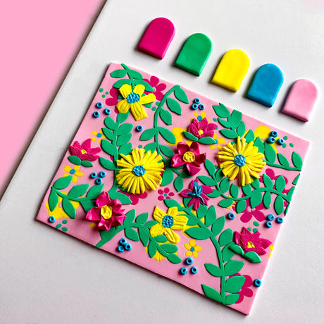 A colourful handmade fimo polymer clay  floral craft plaque on a pink background, decorated with yellow, pink, and blue flowers, green leaves, and small blue bead accents, shown on a white surface with five matching coloured pieces above it.