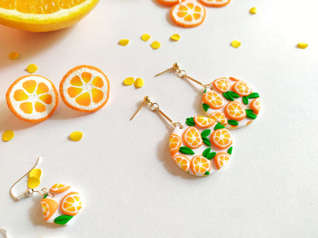 Orange and white polymer clay drop earrings with green leaves, displayed on a white surface with decorative orange slices and yellow pom-pom accents.