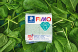 A package of FIMO Effect botanical spinich modelling clay, placed on a bed of fresh green spinach leaves. The packaging is white with blue, red, and green branding, and the clay inside is a pale green speckled shade.
