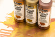 Three bottles of Jacquard Pirata Color Alcohol Ink in Bronze, Rose Gold, and Rose, arranged on a white surface with a gold and orange liquid spill in the foreground.