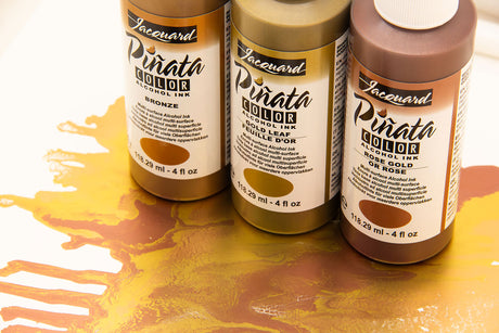 Three bottles of Jacquard Pirata Color Alcohol Ink in Bronze, Rose Gold, and Rose, arranged on a white surface with a gold and orange liquid spill in the foreground.